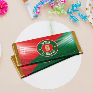 Könnte beinhalten: Zwei Schokoriegel mit rot-grünen Verpackungen, die ein Stadiondesign und den Text "DANIEL 9 IS TURNING" zeigen. Die Riegel haben goldene Folienränder und sind auf einem weißen Kreis platziert, mit Partydekorationen im Hintergrund.