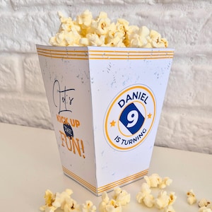 May include: A white popcorn container with orange and blue accents, filled with popcorn. The container has text that reads "Let's Kick Up The Fun!" and "Daniel is turning 9".