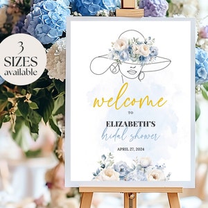 May include: Welcome to Elizabeth's bridal shower sign with a blue and white floral design and a woman's face in a hat. The text is in gold and says "Welcome to Elizabeth's bridal shower, April 27, 2024".