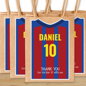 May include: Four brown paper gift bags with a soccer jersey design. The jersey is red and blue with yellow stripes and the name "DANIEL" and number "10" printed on it. The text "THANK YOU for kickin' it with me" is printed on the bottom of the bag.