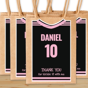 May include: Set of five brown paper gift bags with black and pink jersey-themed thank you tags. The tags feature the name "DANIEL" and the number "10" in pink text. The text "THANK YOU for kickin' it with me" is also included on the tags.
