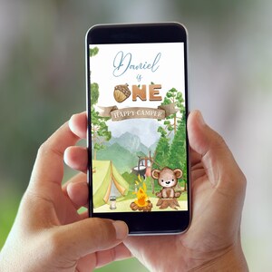 May include: A smartphone displays a watercolor illustration with a camping theme. The design includes the text "Daniel is ONE HAPPY CAMPER" with a bear, tent, campfire, and forest backdrop. The overall color palette is soft and inviting.