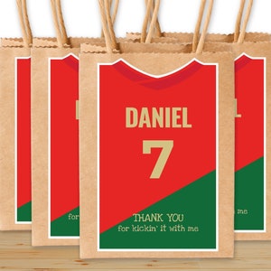 Peut inclure: Ensemble de sacs cadeaux en papier kraft avec des étiquettes à thème de maillot de football rouge et vert. Les étiquettes présentent le nom "DANIEL" et le numéro "7" en lettres dorées. Le texte "MERCI d'avoir joué avec moi" est imprimé sur les étiquettes.