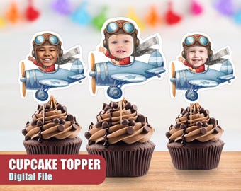 May include: Three cupcake toppers featuring a cartoon airplane with a child's face in the cockpit. The airplanes are blue and white with red accents. The toppers are perfect for a birthday party.