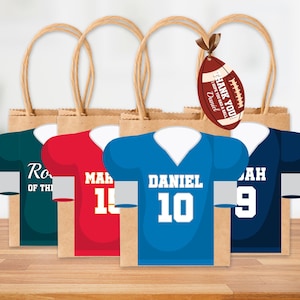 May include: Four brown paper gift bags with football jersey designs in blue, red, and green. Each bag has a jersey shape with a name and number. A football-shaped tag says "Thank You! I hope you had a ball, Daniel."