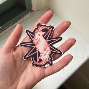 May include: A sticker featuring a pink goat-like creature with horns, set against a black starburst design. The sticker has a white border and is held in a hand. The overall design has a whimsical, mystical theme.