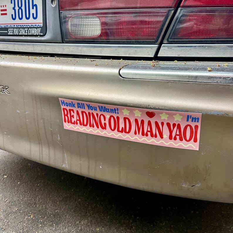 May include: A pink bumper sticker with the text "Honk All You Want! I'm Reading Old Man Yaoi" in red and white letters. There are three yellow stars and a red heart on the sticker.