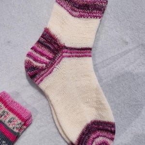 May include: A hand-knitted sock in cream, with a cuff and heel in shades of purple and pink. The sock features horizontal stripes in varying shades of purple and pink. The other sock is partially visible.