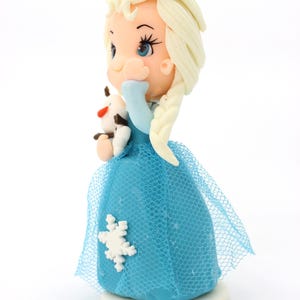 May include: A figurine of a character with blonde hair in a side braid, wearing a blue dress with a white snowflake accent and a blue tulle overlay. The character holds a small white snowman figure with a carrot nose and brown stick arms.