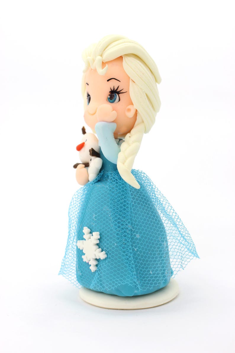 May include: A figurine of a character with blonde hair in a side braid, wearing a blue dress with a white snowflake accent and a blue tulle overlay. The character holds a small white snowman figure with a carrot nose and brown stick arms.