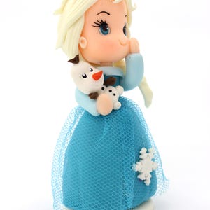 May include: A small figurine of a character with pale blonde hair, blue eyes, and a light blue dress with a blue tulle overlay. The character holds a small white snowman figure with a carrot nose and black buttons, featuring a white snowflake accent.