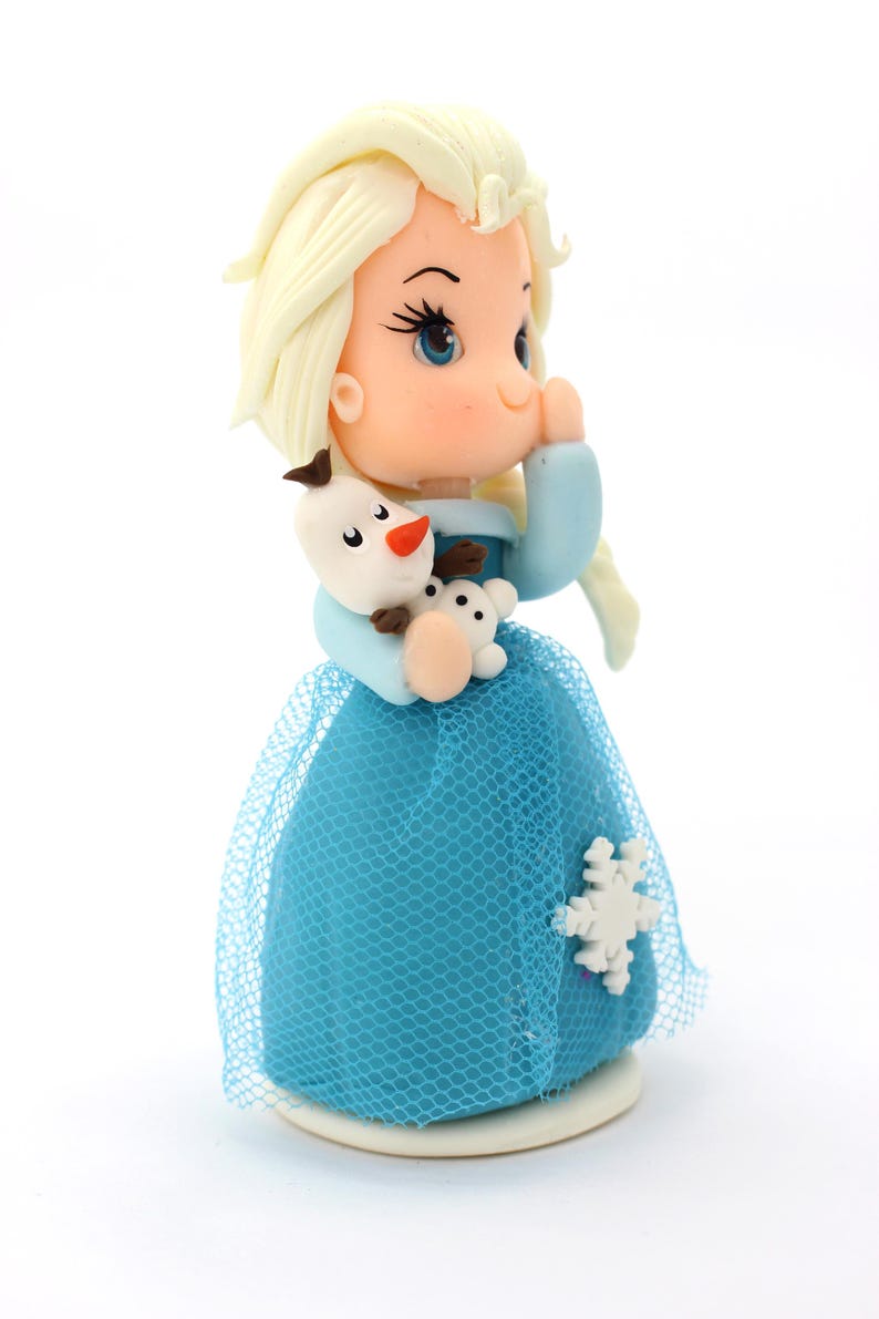 May include: A small figurine of a character with pale blonde hair, blue eyes, and a light blue dress with a blue tulle overlay. The character holds a small white snowman figure with a carrot nose and black buttons, featuring a white snowflake accent.