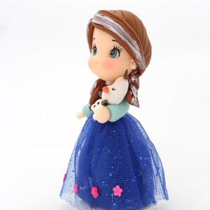 May include: A figurine of a character with brown hair featuring a white streak, wearing a light blue top and a dark blue glittery tulle skirt adorned with small pink flower accents. The character holds a small white snowman figure with an orange nose.