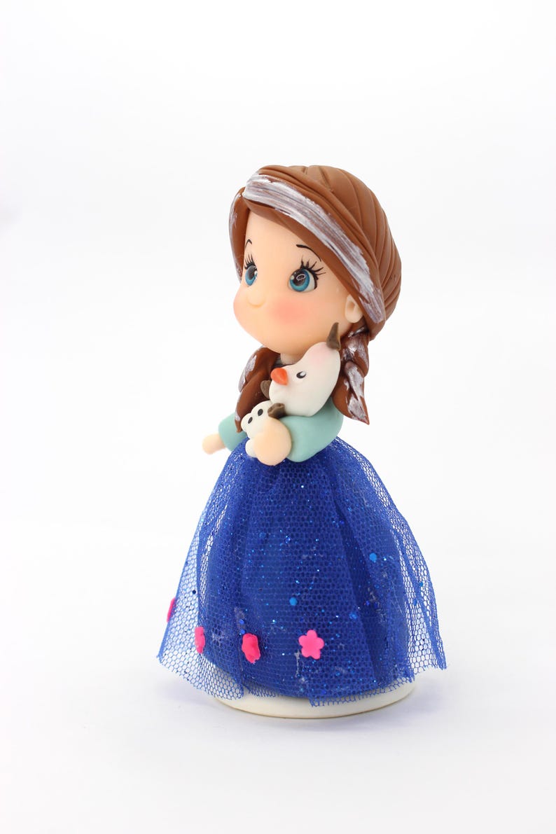 May include: A figurine of a character with brown hair featuring a white streak, wearing a light blue top and a dark blue glittery tulle skirt adorned with small pink flower accents. The character holds a small white snowman figure with an orange nose.