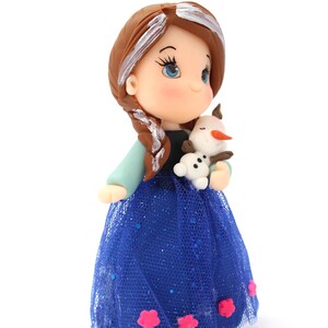 May include: A figurine of a character with brown hair in a side braid, wearing a black bodice and a blue tulle skirt with pink flower accents. The character holds a small white snowman figure with an orange nose and brown twig arms.