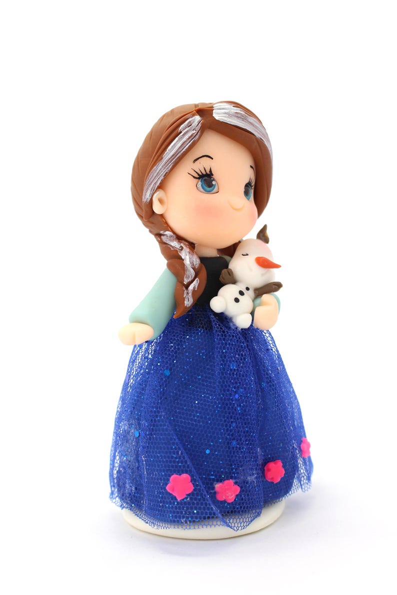 May include: A figurine of a character with brown hair in a side braid, wearing a black bodice and a blue tulle skirt with pink flower accents. The character holds a small white snowman figure with an orange nose and brown twig arms.