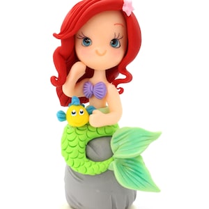 May include: A figurine of a mermaid with vibrant red hair, a small pink star hair accessory, and a purple seashell top. The mermaid has a bright green tail and is perched on a gray rock, holding a small yellow and blue fish character.
