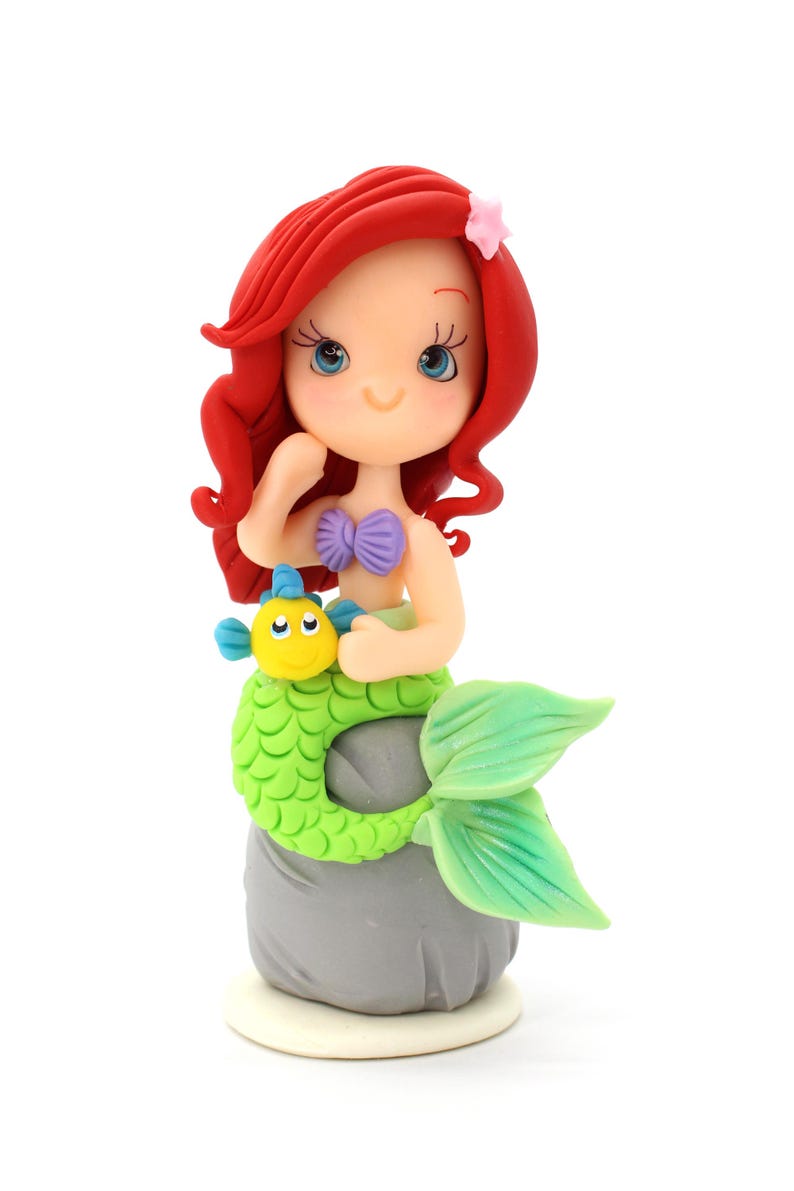 May include: A figurine of a mermaid with vibrant red hair, a small pink star hair accessory, and a purple seashell top. The mermaid has a bright green tail and is perched on a gray rock, holding a small yellow and blue fish character.