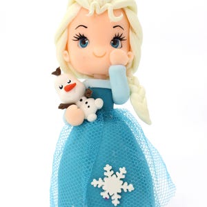May include: A figurine of a character with pale skin, blue eyes, and long blonde hair in a braid, wearing a blue dress with a sheer blue tulle overlay and a white snowflake accent, holding a small white snowman figure with a carrot nose.