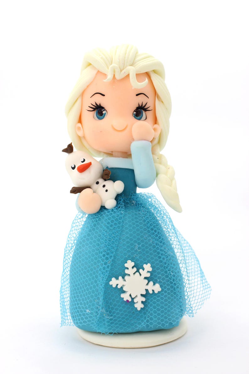 May include: A figurine of a character with pale skin, blue eyes, and long blonde hair in a braid, wearing a blue dress with a sheer blue tulle overlay and a white snowflake accent, holding a small white snowman figure with a carrot nose.