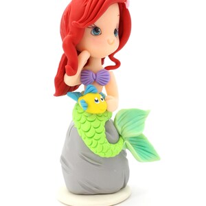 May include: A figurine of a mermaid with long red hair, a purple seashell top, and a green scaly tail, sitting on a gray rock. A small yellow and blue fish is positioned in front of the mermaid. The figure has blue eyes and a small pink flower.
