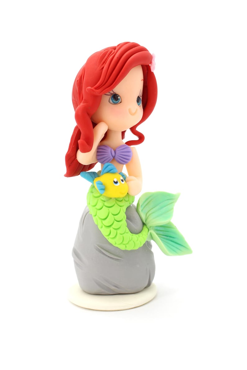 May include: A figurine of a mermaid with long red hair, a purple seashell top, and a green scaly tail, sitting on a gray rock. A small yellow and blue fish is positioned in front of the mermaid. The figure has blue eyes and a small pink flower.