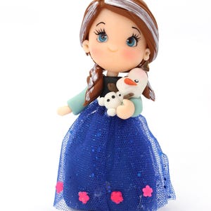 May include: A figurine of a character with brown hair featuring white highlights, blue eyes, and rosy cheeks. The character wears a black top with light blue sleeves and a long, glittery blue tulle skirt adorned with small pink flower accents.
