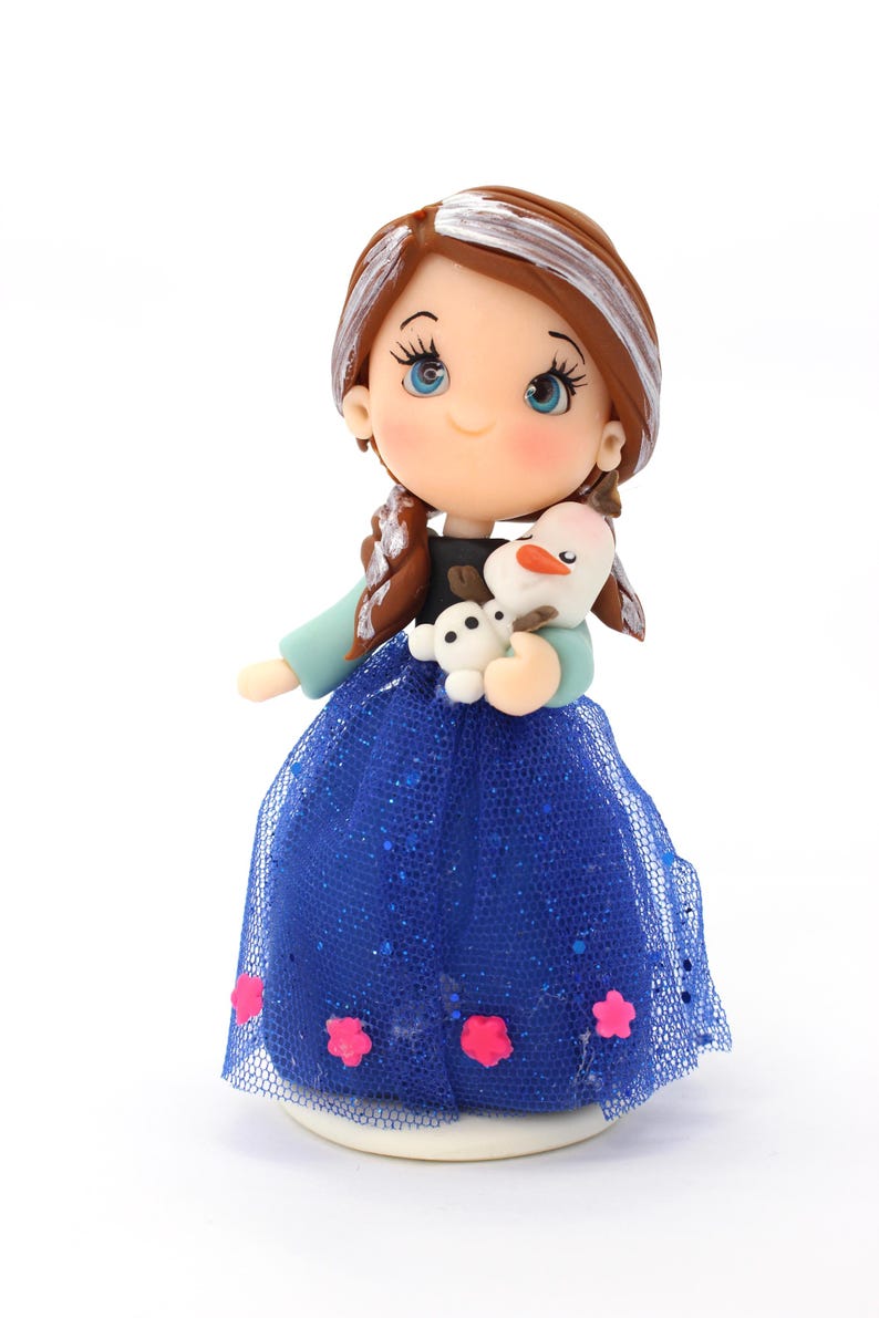 May include: A figurine of a character with brown hair featuring white highlights, blue eyes, and rosy cheeks. The character wears a black top with light blue sleeves and a long, glittery blue tulle skirt adorned with small pink flower accents.