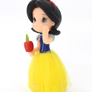 May include: A small figurine of a character with black hair, a red bow, and blue bodice, wearing a yellow tulle skirt with glitter accents. The figure holds a small red apple with a green leaf in one hand and has a hand raised to the face.