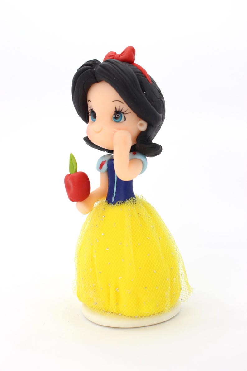 May include: A small figurine of a character with black hair, a red bow, and blue bodice, wearing a yellow tulle skirt with glitter accents. The figure holds a small red apple with a green leaf in one hand and has a hand raised to the face.
