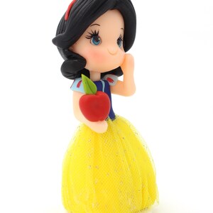 May include: A figurine of a character with black hair, a red headband, and blue bodice, wearing a yellow tulle skirt with glitter accents. The character holds a red apple with a green leaf in one hand and has one hand raised to the cheek.