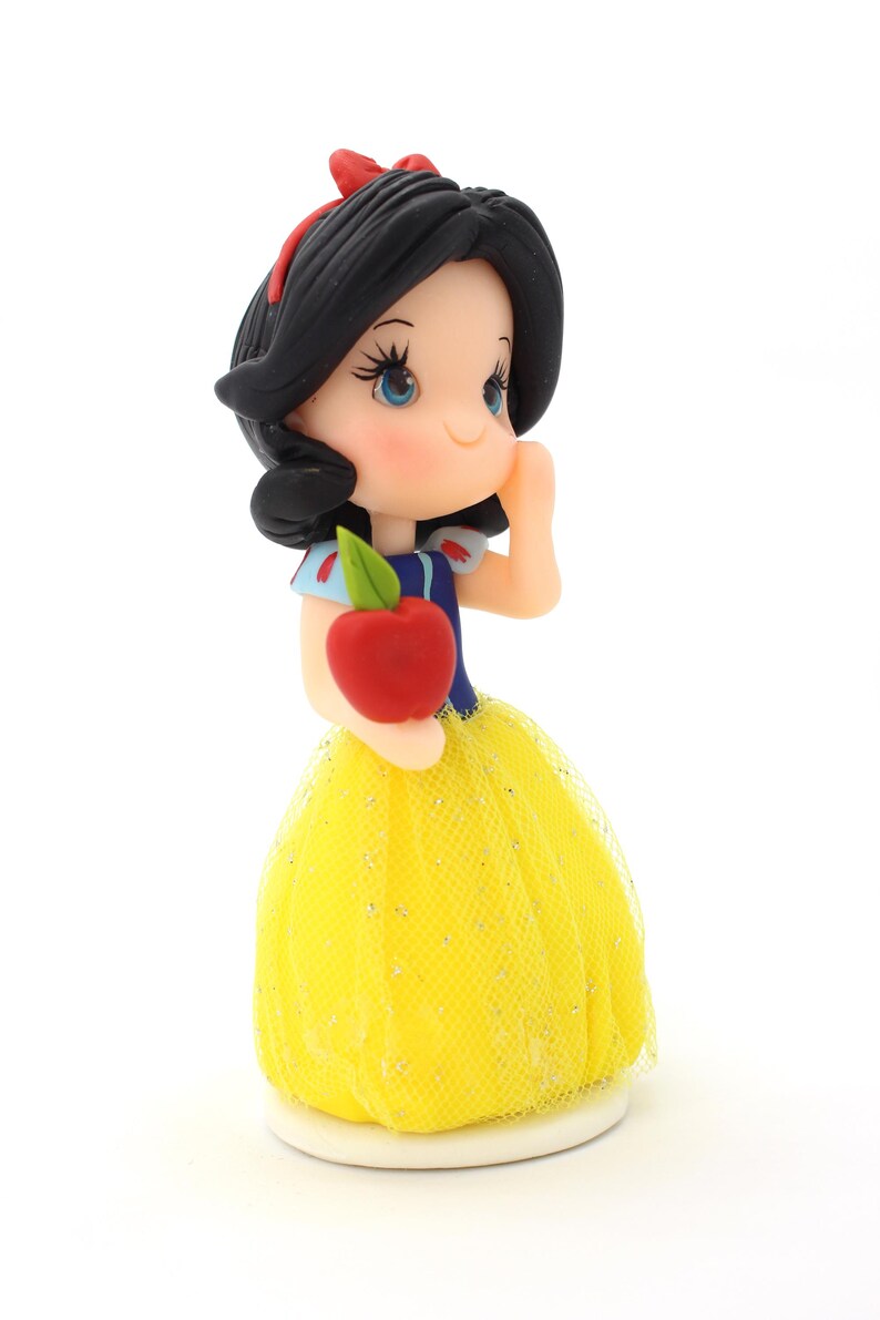 May include: A figurine of a character with black hair, a red headband, and blue bodice, wearing a yellow tulle skirt with glitter accents. The character holds a red apple with a green leaf in one hand and has one hand raised to the cheek.