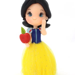 May include: A figurine of a character with black hair, a red bow, and blue eyes, wearing a blue bodice with puffed sleeves and a yellow tulle skirt with silver glitter. The character holds a red apple with a green leaf in one hand.