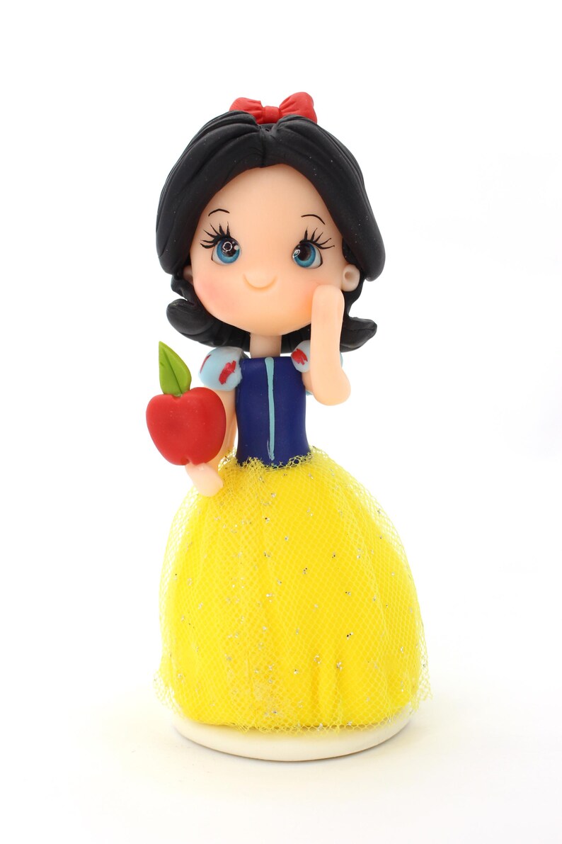 May include: A figurine of a character with black hair, a red bow, and blue eyes, wearing a blue bodice with puffed sleeves and a yellow tulle skirt with silver glitter. The character holds a red apple with a green leaf in one hand.
