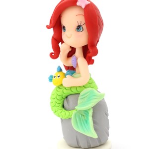 May include: A figurine of a mermaid with vibrant red hair, a pale complexion, and a bright green scaly tail. The mermaid sits on a gray rock, holding a small yellow and blue fish. A light pink starfish is attached to the red hair.