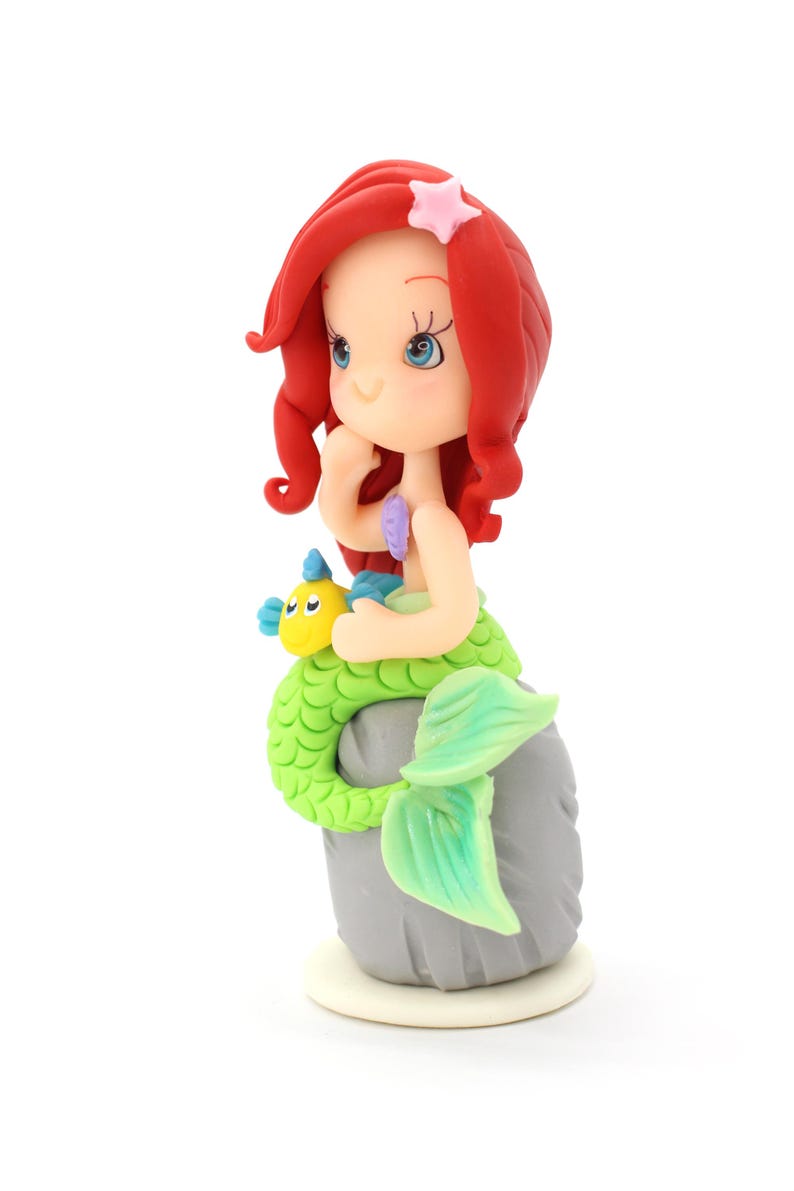 May include: A figurine of a mermaid with vibrant red hair, a pale complexion, and a bright green scaly tail. The mermaid sits on a gray rock, holding a small yellow and blue fish. A light pink starfish is attached to the red hair.