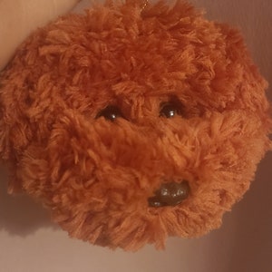 May include: A brown plush toy shaped like a dog with black eyes and a brown nose. The toy is hanging from a gold chain.