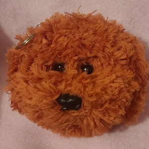 May include: A plush brown dog keyring with black eyes and a black nose. The keyring is made of soft, fluffy material.