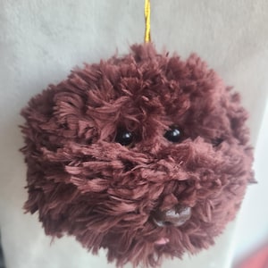 May include: A brown plush toy shaped like a dog's head with black eyes and a pink tongue. The toy is hanging from a gold string.