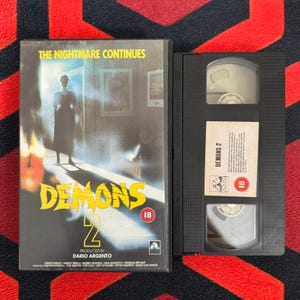 Könnte beinhalten: Eine Vintage-VHS-Kassette des Horrorfilms "Demons 2". Das Cover zeigt eine schattenhafte Figur, Flammen und den Filmtitel in Gelb. Die Kassette ist schwarz, mit Titel und Bewertung sichtbar.