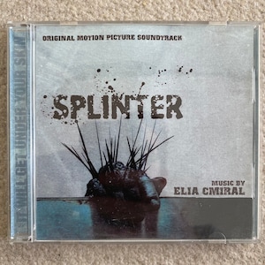 Könnte beinhalten: Ein CD-Gehäuse mit grauem Hintergrund und dem Text "SPLINTER" in Schwarz. Der Text "ORIGINAL MOTION PICTURE SOUNDTRACK" ist ebenfalls in Schwarz. Das Bild zeigt eine Hand, die aus dem Wasser ragt, mit einer Pflanze, die aus ihr herauswächst. Der Text "MUSIC BY ELIA CMIRAL" ist in Schwarz in der unteren rechten Ecke.