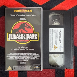 May include: A Jurassic Park VHS tape and its black plastic case. The cover features a red circle with a black dinosaur silhouette, the title text, and the phrase An Adventure 65 Million Years In The Making. The tape has a white label with text.