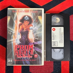 Peut inclure: Une cassette VHS de Prom Night II Hello Mary Lou avec un boîtier blanc montrant une femme en robe violette et diadème. Le texte indique Mary-Lou is back...again!, Prom Night II Hello Mary Lou, 18, et Vengeance never rests in peace. Sheer Entertainment.
