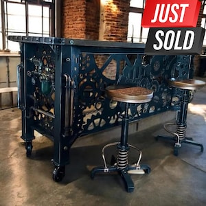 May include: A black metal bar table with a gear design and three black metal bar stools with a spring mechanism. The bar table has wheels and is made of metal. The bar stools have a wooden seat. The bar table and stools are in a rustic industrial style.
