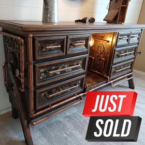 May include: A dark wood dresser with copper pipe accents and steampunk-inspired details. The dresser has six drawers with brass handles and a large open compartment in the center. The drawers are decorated with gears and other steampunk elements. The dresser is lit from within, casting a warm glow. The text "JUST SOLD" is superimposed on the image.