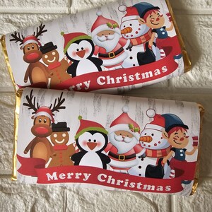 May include: A white chocolate bar with a festive Christmas design. The bar features a reindeer, gingerbread man, penguin, Santa Claus, snowman, and elf. The text "Merry Christmas" is printed on a red ribbon.