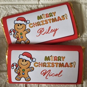 Może przedstawiać: Dwie czekolady owinięte w złotą folię, każda z czerwoną etykietą. Etykiety przedstawiają piernikowego ludzika w czapce Świętego Mikołaja, napisy "MERRY CHRISTMAS!" i imię. Jeden mówi "Riley", a drugi "Nicol".