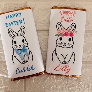 Puede incluir: Dos envoltorios de barritas de chocolate con ilustraciones de conejos. Un envoltorio es azul y dice "¡Feliz Pascua! Carter". El otro envoltorio es rosa y dice "Feliz Pascua Lilly".