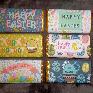 May include: Six chocolate bars wrapped in foil with different Easter-themed designs. The designs feature bunnies, eggs, flowers, and the text "Happy Easter".