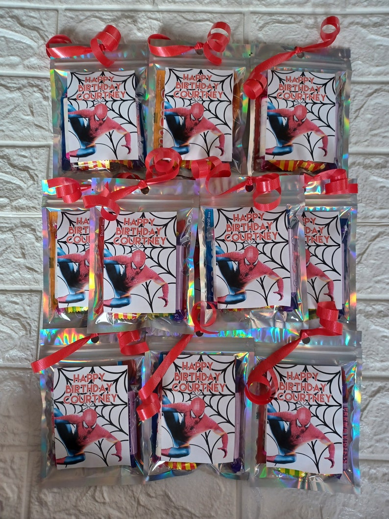 May include: Nine small clear bags with red ribbon ties, each containing a variety of colorful items and a printed card with the text "Happy Birthday Courtney" and a cartoon image of Spiderman.